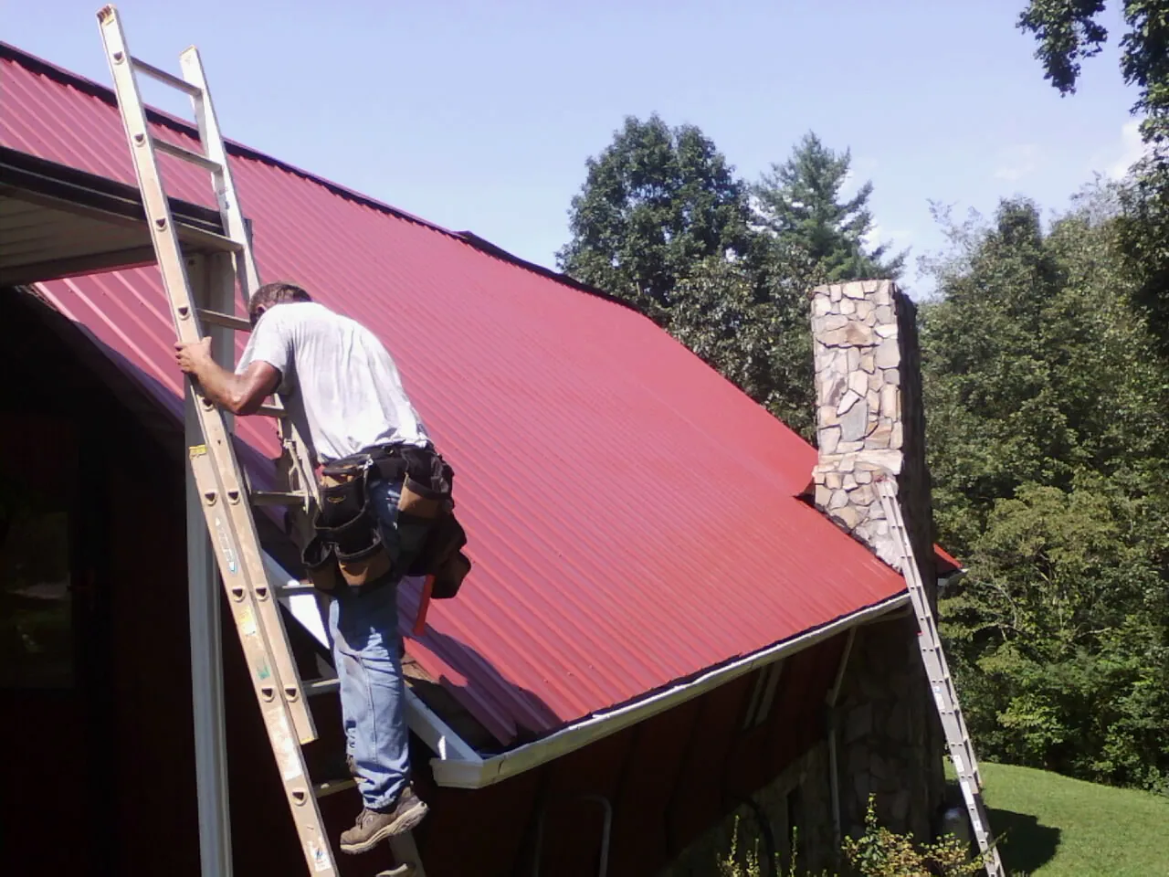 Roofer on a ladder beside a red metal roof and stone chimney — Soffit & Fascia Repair by Roofing Brothers in Maryland Heights